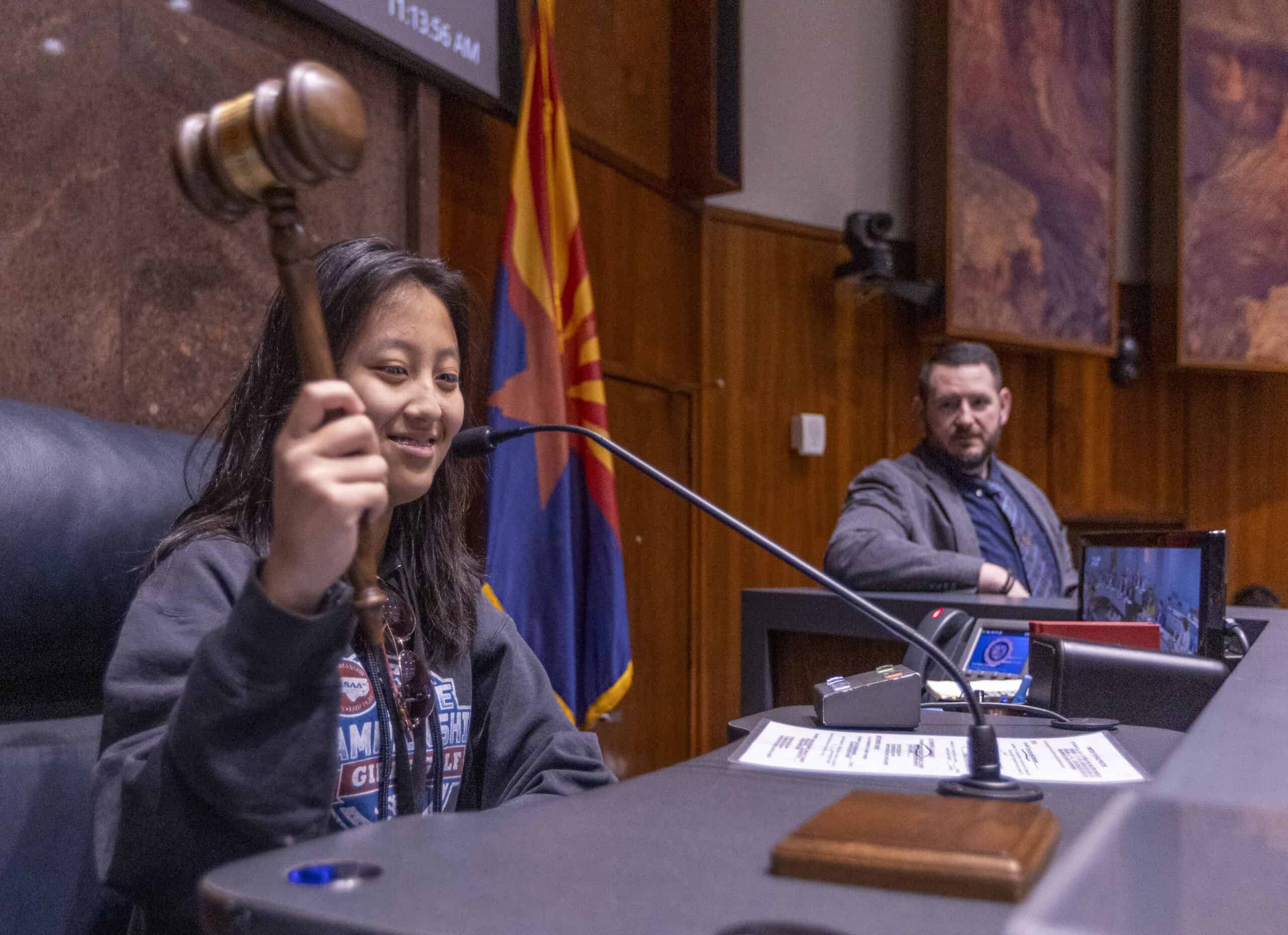 A young person smiles while holding a gavel at a judge’s desk in a chamber. The Citizen watches from the background as the Arizona state flag hangs between them, with papers and microphones on the desk.