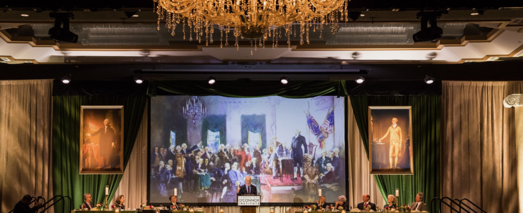 A grand banquet hall with a large chandelier, featuring a speaker at a podium. A painting depicting a historical scene is displayed on a screen behind them. Portraits of historical figures hang on either side, and guests are seated at a long table.