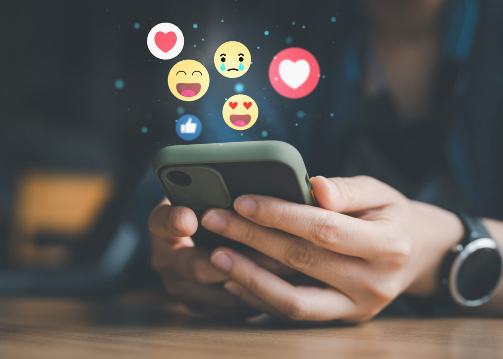 A person holds a smartphone with both hands. Floating emojis, including a heart, smiley face, crying face, and thumbs-up, appear above the phone, indicating social media interactions. The background is blurred.