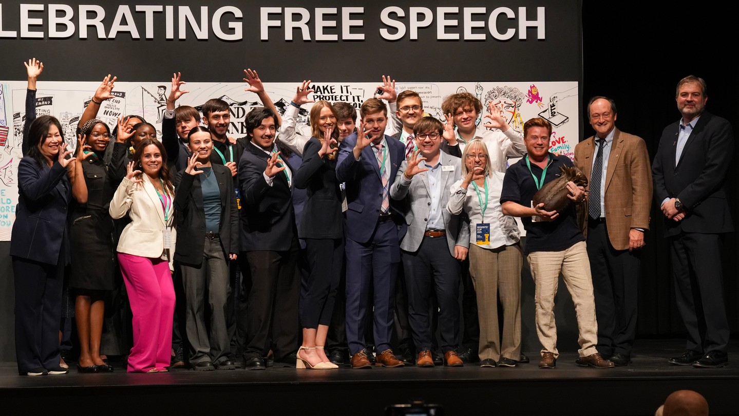 491446585_18366395893132731_3025261584145495845_n A diverse group of people poses on a stage under a sign that reads "CELEBRATING FREE SPEECH." Many are smiling and making an L-shaped hand gesture, with illustrated posters in the background.