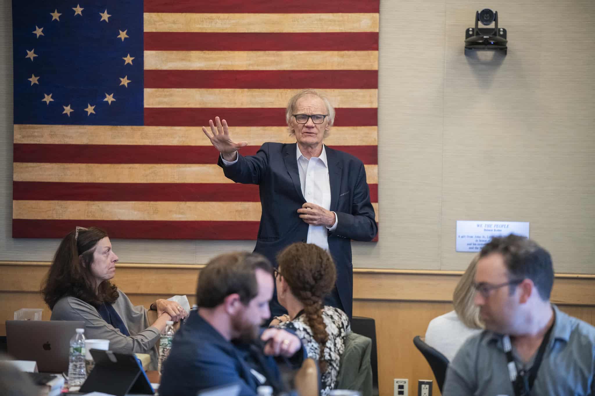 A man in a suit gestures while speaking to a seated audience in a classroom, engaging them in Civics Education. Behind him is a historic American flag on the wall. Several people are seated at tables with laptops and water bottles.