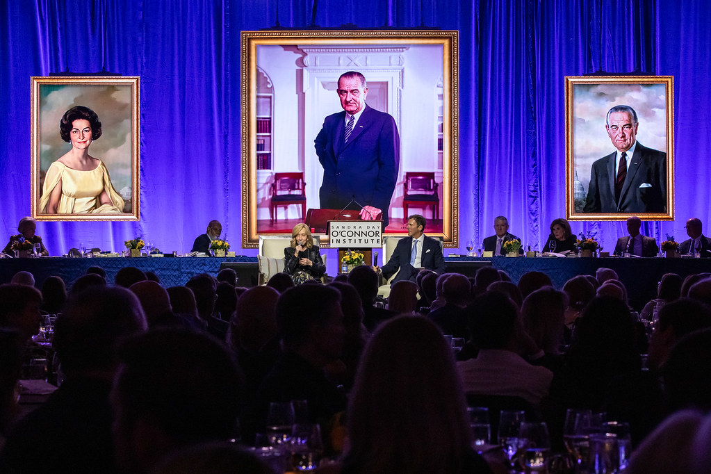 A formal event with many attendees features two people, including Doris Kearns Goodwin, in discussion onstage. Behind them is a blue curtain and large portraits of two individuals on either side, with a larger central portrait of a man in a suit.