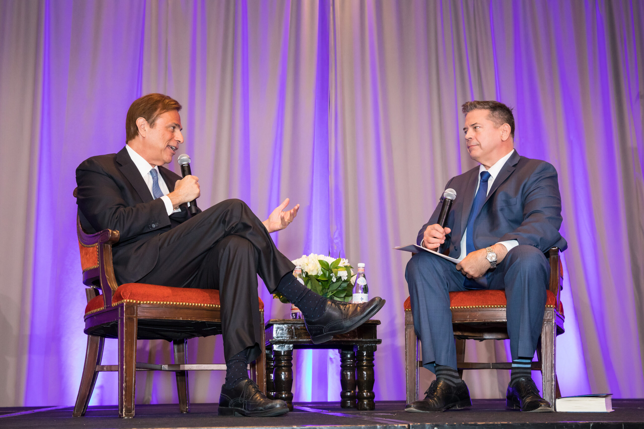 Two men in suits are seated on a stage, engaging in conversation while holding microphones. They are positioned in front of a purple curtain with a small table and a flower arrangement between them.