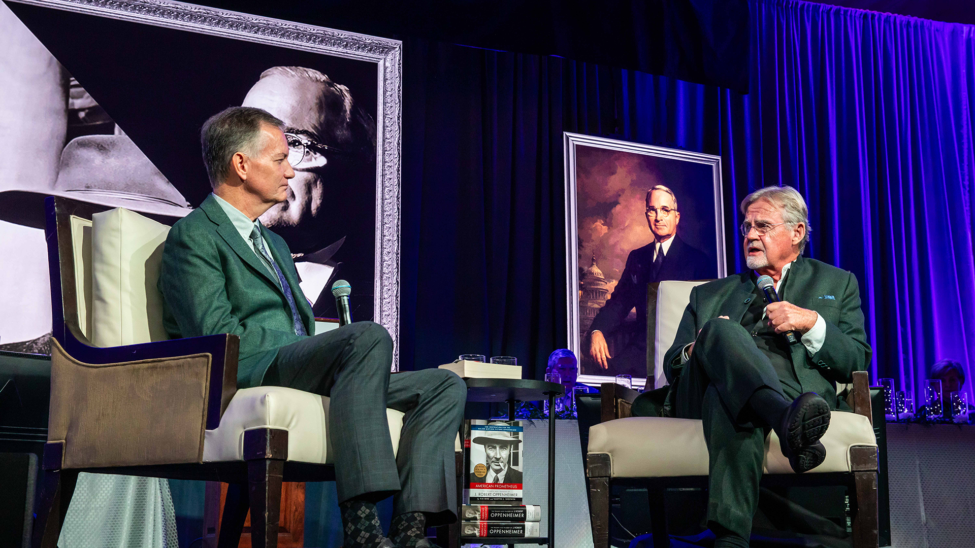 Two men in suits sit on stage holding microphones, engaged in discussion. Behind them are portraits of different individuals. A small table with books is placed between the chairs. The setting is richly lit with blue and purple tones.