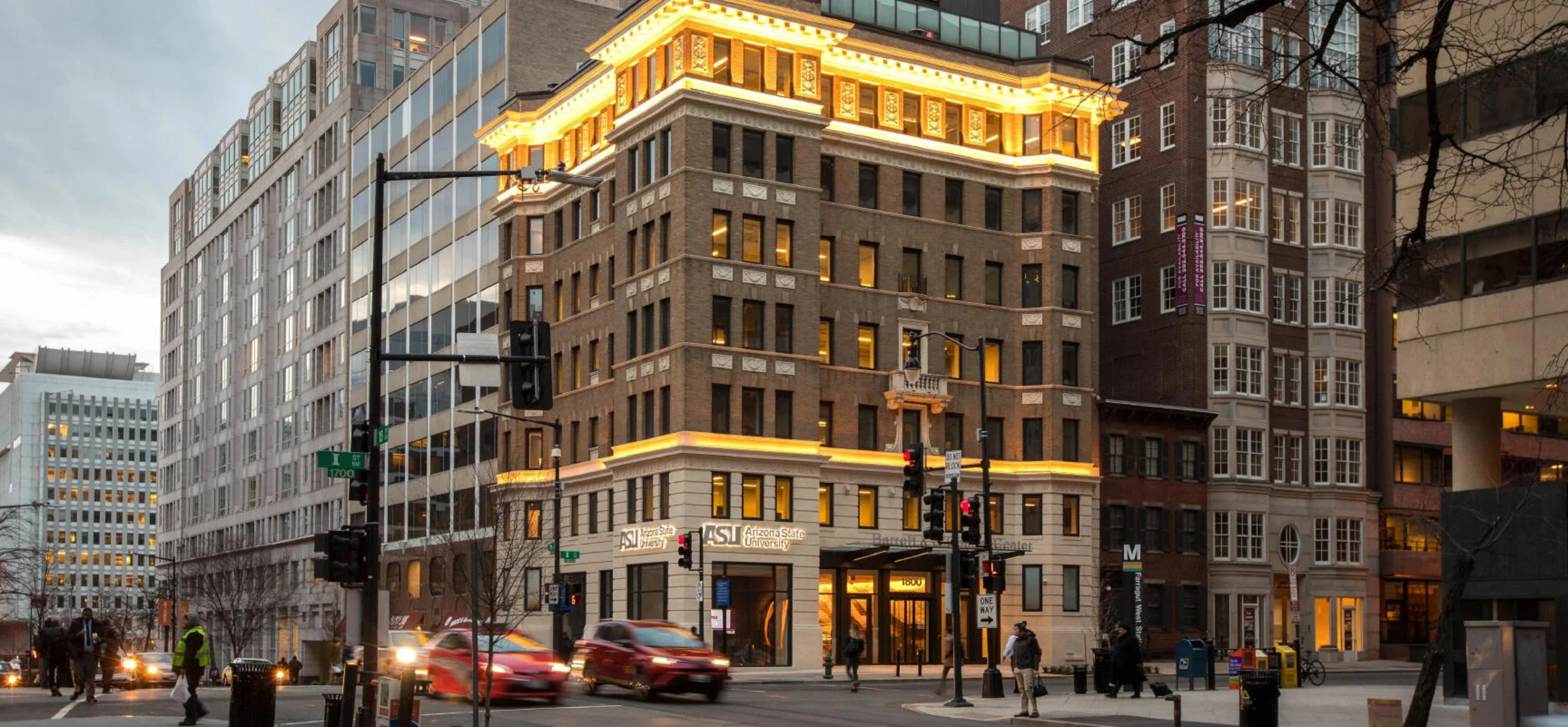 A city street at dusk in Washington D.C., with cars driving by. A historic multi-story building with illuminated trim stands at the corner, surrounded by modern and older buildings, as Washington D.C. Fellowship members wait at the crosswalk.