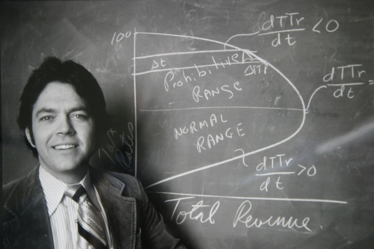About-Dr-Laffer A man in a suit stands smiling beside a chalkboard with economic graphs and terms like "Prohibitive Range," "Normal Range," and "Total Revenue" written on it. Mathematical equations are also visible on the board.