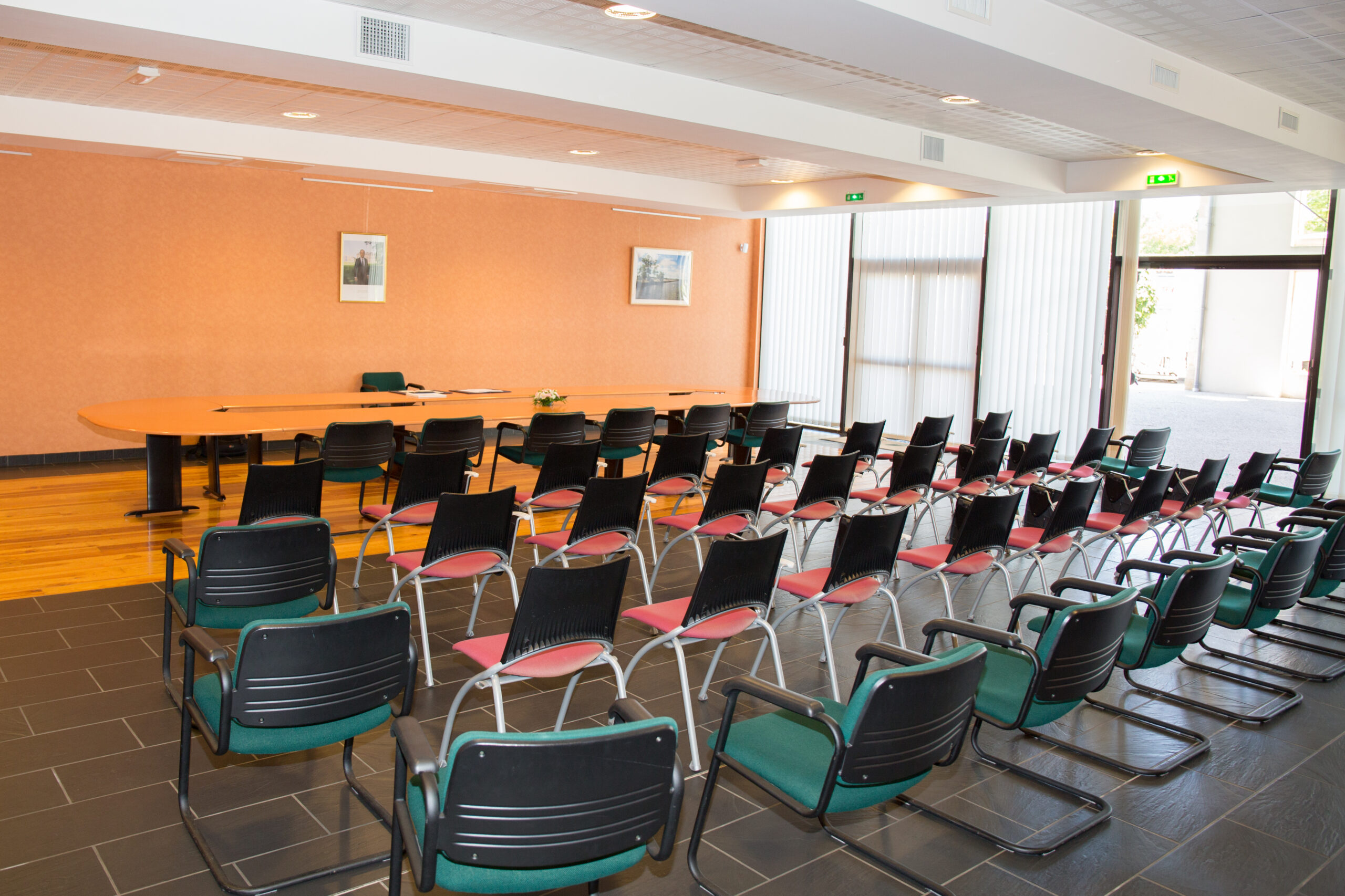 Large Room with chair in a city hall of village A conference room with rows of empty chairs faces a long table adorned with documents and flowers, ready for the Summer of Civics gathering, beneath a peach-colored wall with framed pictures and bathed in natural light from large windows.