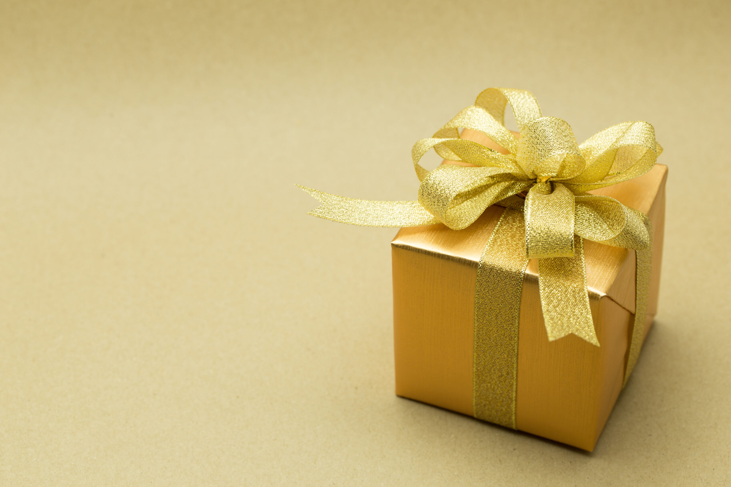 A small gold gift box with a shiny gold ribbon and bow, set against a plain beige background.