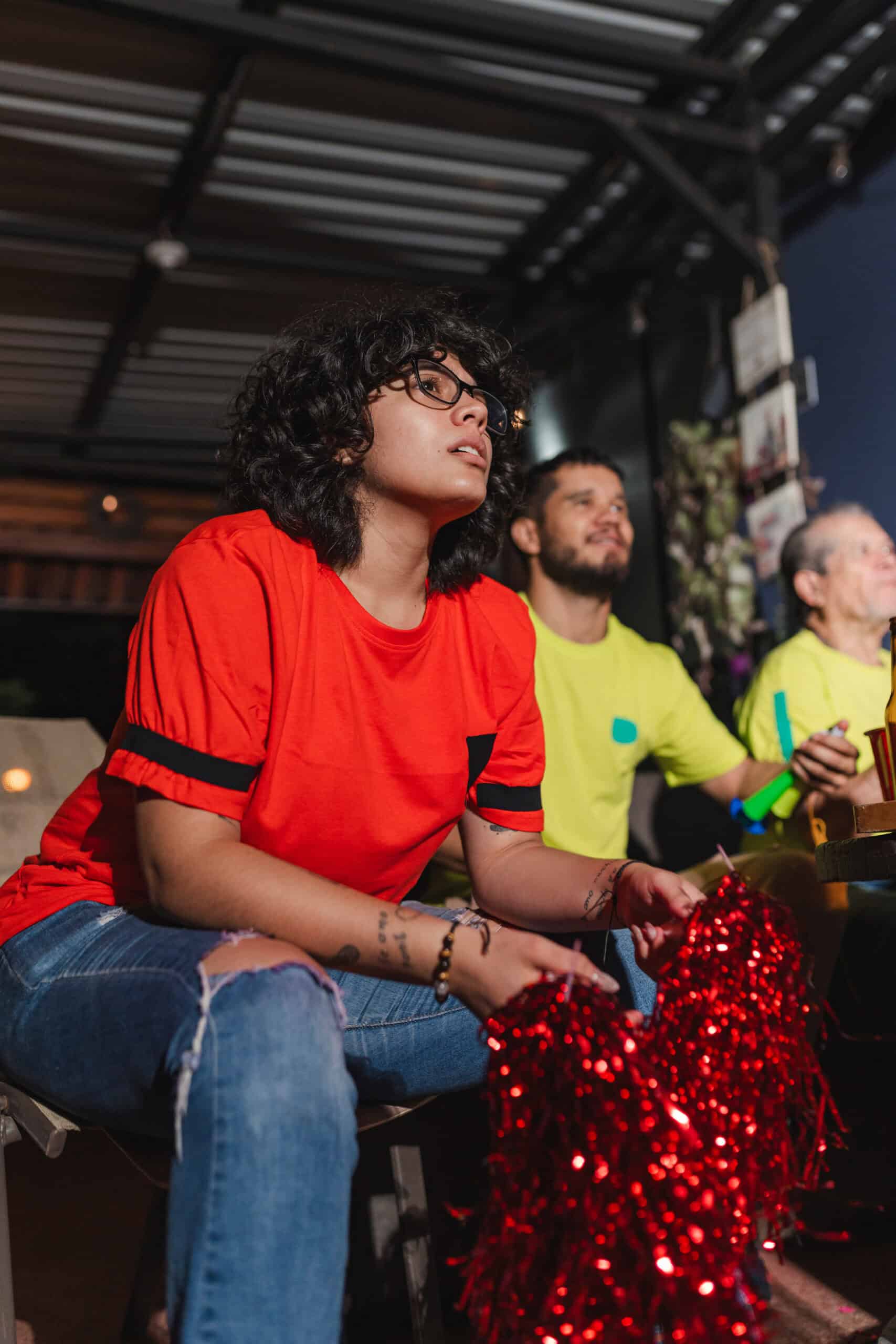 A person in a red shirt with curly hair and glasses holds red pom-poms, looking focused—perhaps supporting their team at a World Cup viewing. Two others, one in yellow, sit nearby as they all watch the event unfold indoors.