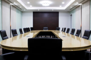 A modern, empty conference room with a large, U-shaped wooden table surrounded by black chairs. The room has bright overhead lighting, white walls, and a dark accent wall at the far end.