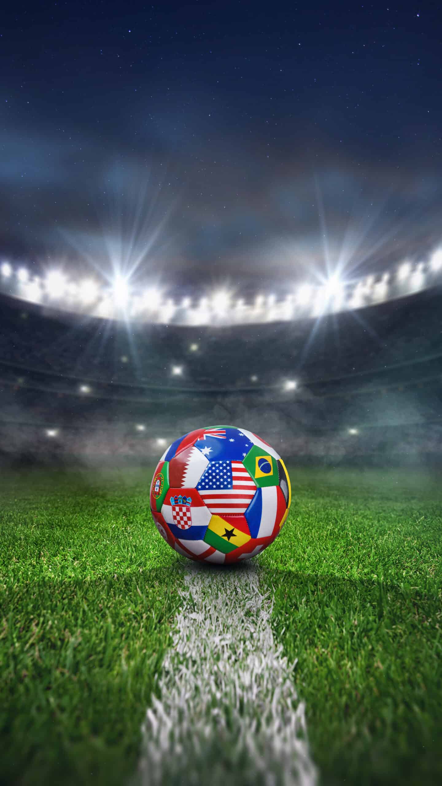 A soccer ball with world flags sits on a white line on a green field in a brightly lit stadium at night, commemorating America’s 250th Anniversary.