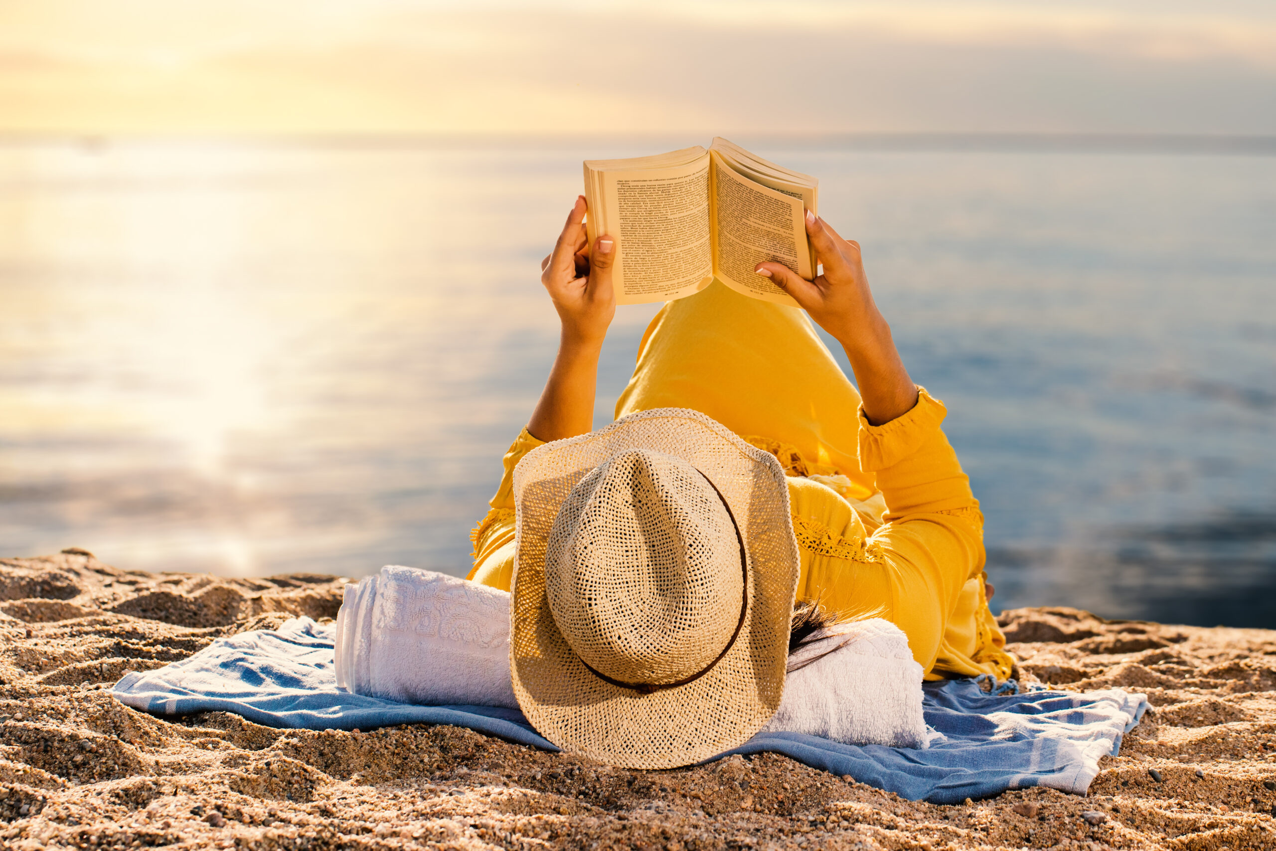 Woman reading a novel at sunset on beach. A person in a yellow outfit lies on a beach towel by the sea at sunset, holding an open book above their face—a perfect summer of civics moment. A straw hat rests on their chest as the softly lit sky sets the scene.