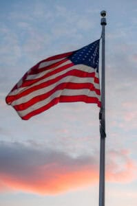 The American flag waves on a flagpole against a colorful sky at sunset, evoking the spirit of a Summer of Civics with soft clouds tinted in shades of pink, orange, and blue.