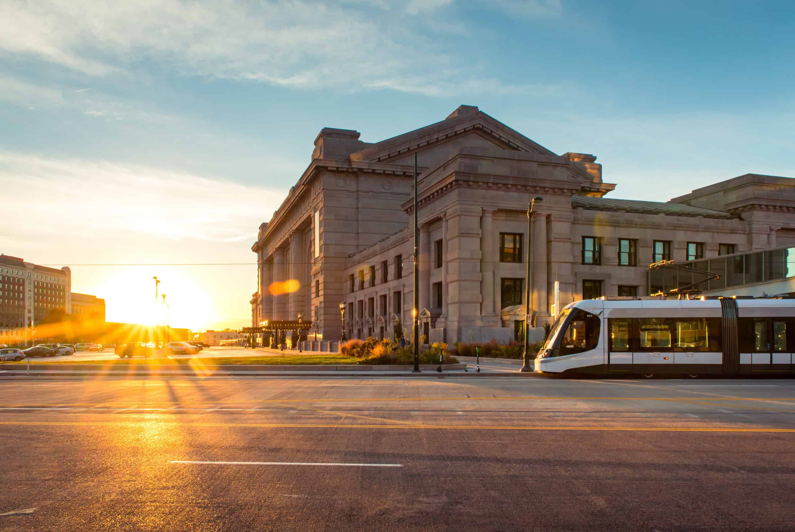 A modern streetcar passes in front of a large, historic stone building at sunset, with sunlight casting long shadows across the street as banners celebrating the World Cup flutter in the warm evening breeze.