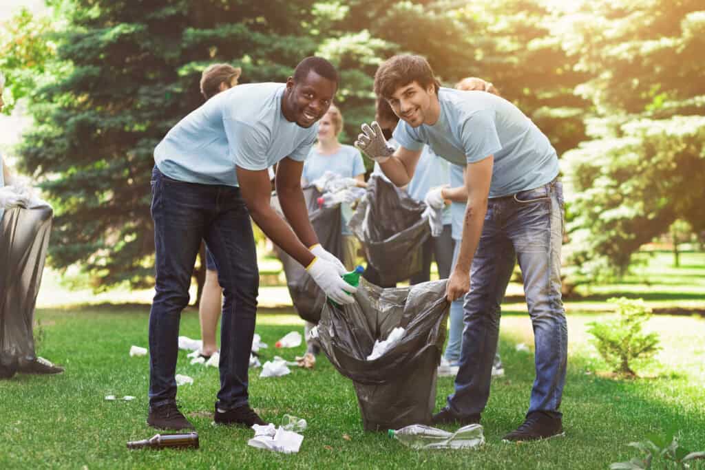 Two young men wearing gloves and light blue shirts pick up litter as ambassadors for the environment, smiling at the camera. Other volunteers are cleaning up in the background among trees and grass.