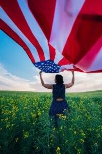 A person standing in a field of yellow flowers on a bright spring day holds up a large American flag, with the flag billowing above them against a blue sky with scattered clouds.