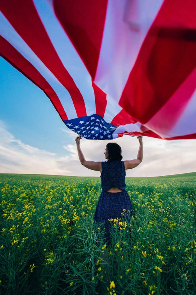 A person standing in a field of yellow flowers on a bright spring day holds up a large American flag, with the flag billowing above them against a blue sky with scattered clouds.