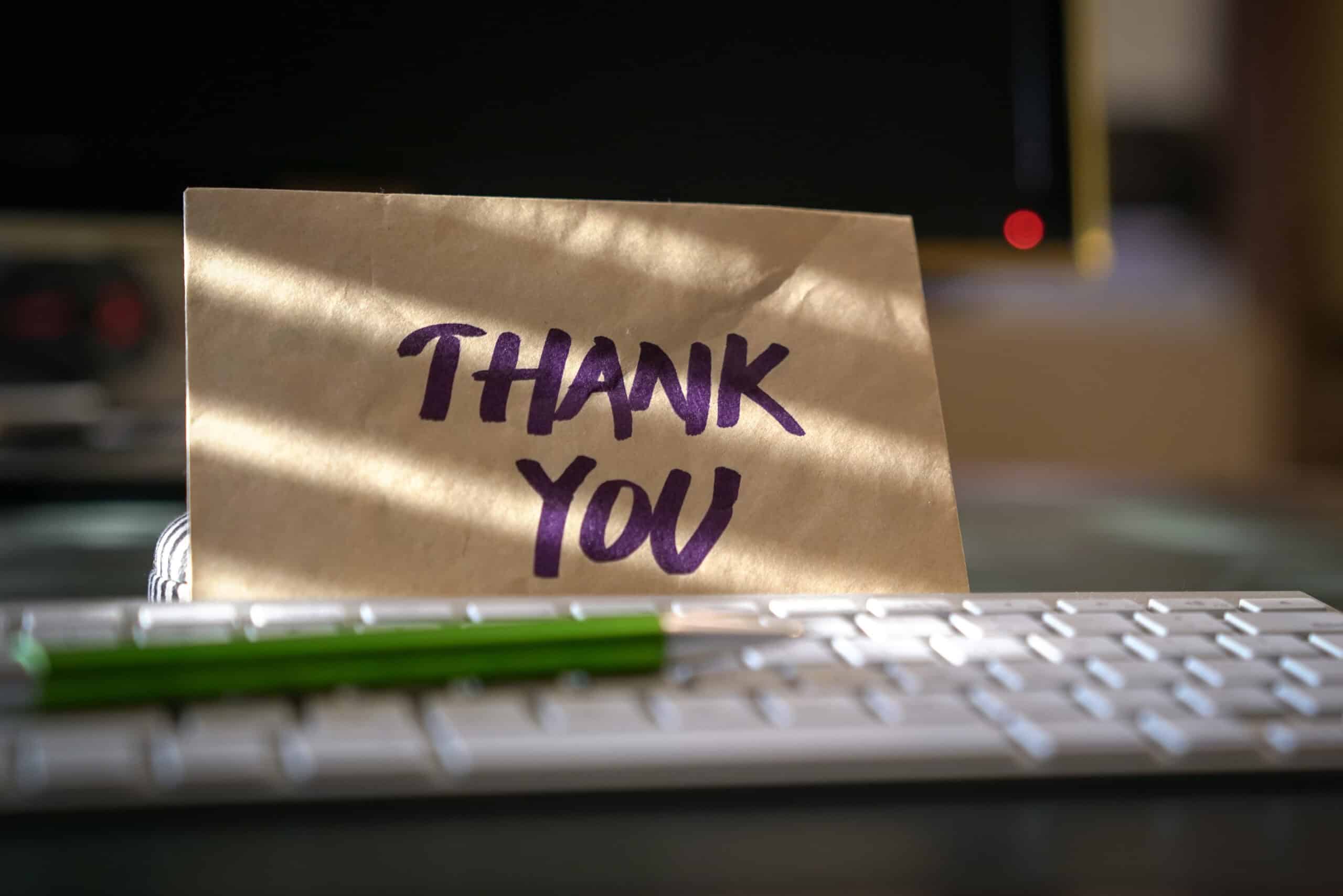 A brown card with "THANK YOU" in bold purple letters is propped up behind a white computer keyboard. A green pen rests on the keyboard, while sunlight and hints of spring cast gentle shadows across the scene.