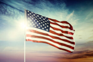 The American flag waves in the wind against a vibrant sky at sunrise or sunset, with sunlight shining through—capturing the spirit of America 250 and our nation’s enduring legacy.