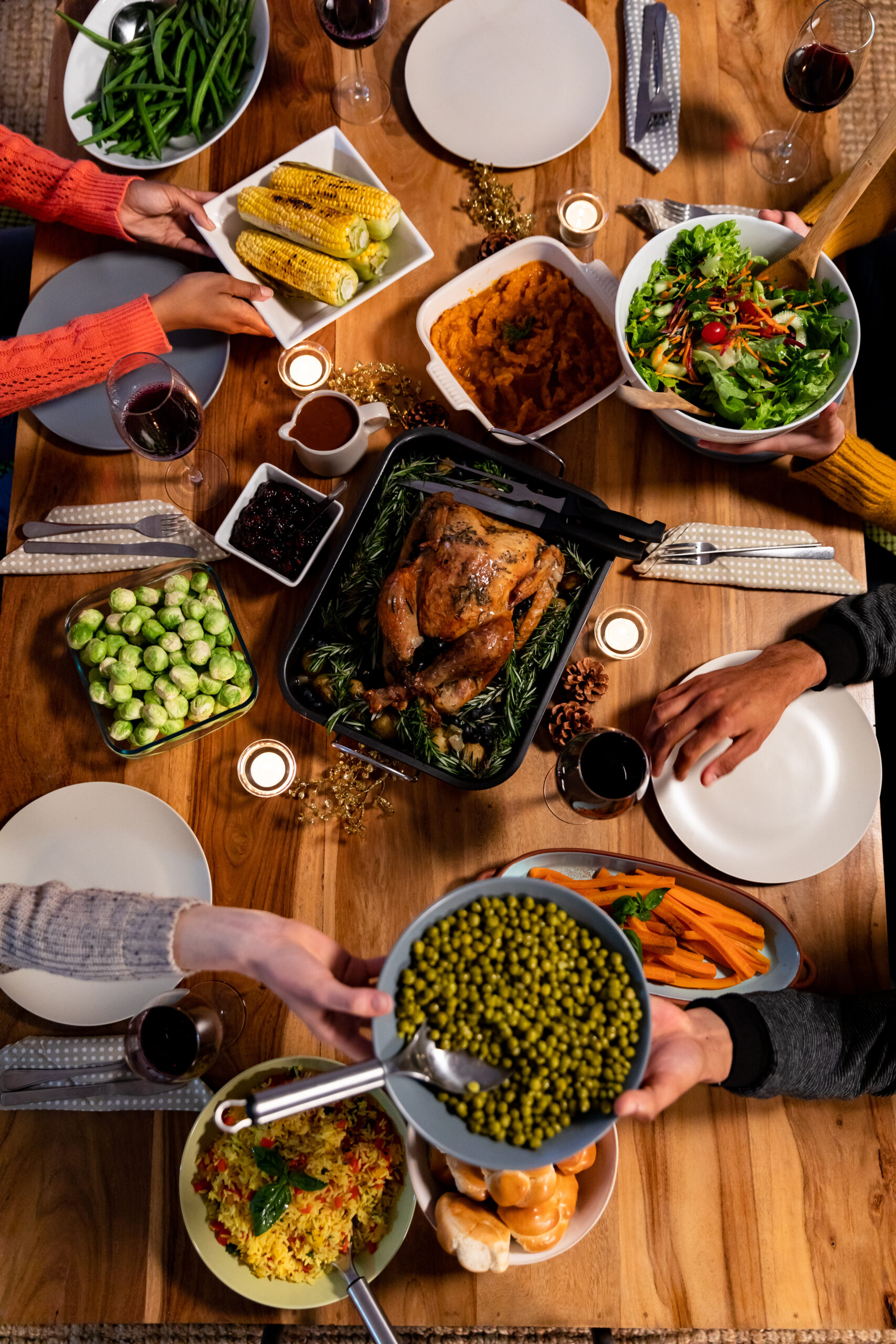 A dinner table set for a festive meal reminiscent of America's Founders, featuring roast chicken, mashed sweet potatoes, corn on the cob, green beans, Brussels sprouts, salad, carrots, bread rolls, candles, and people serving food.