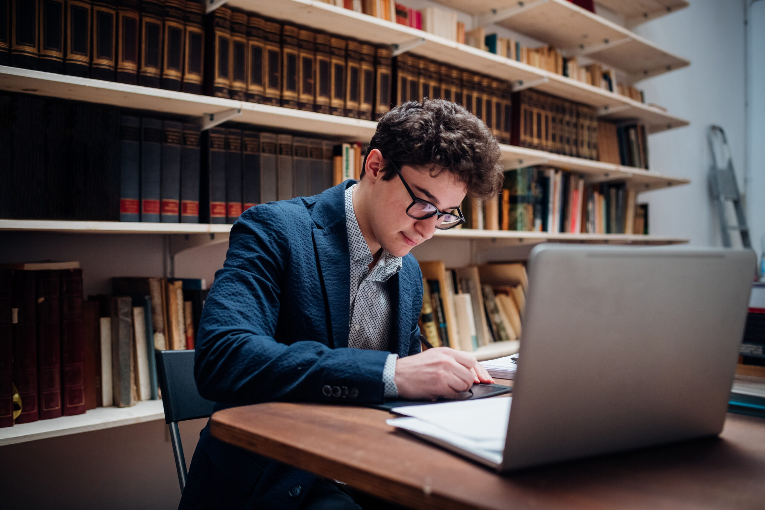 A young man with curly hair and glasses studies at a table in a library in Washington D.C., looking at papers in front of a laptop, surrounded by bookshelves, as he prepares for his prestigious Fellowship.