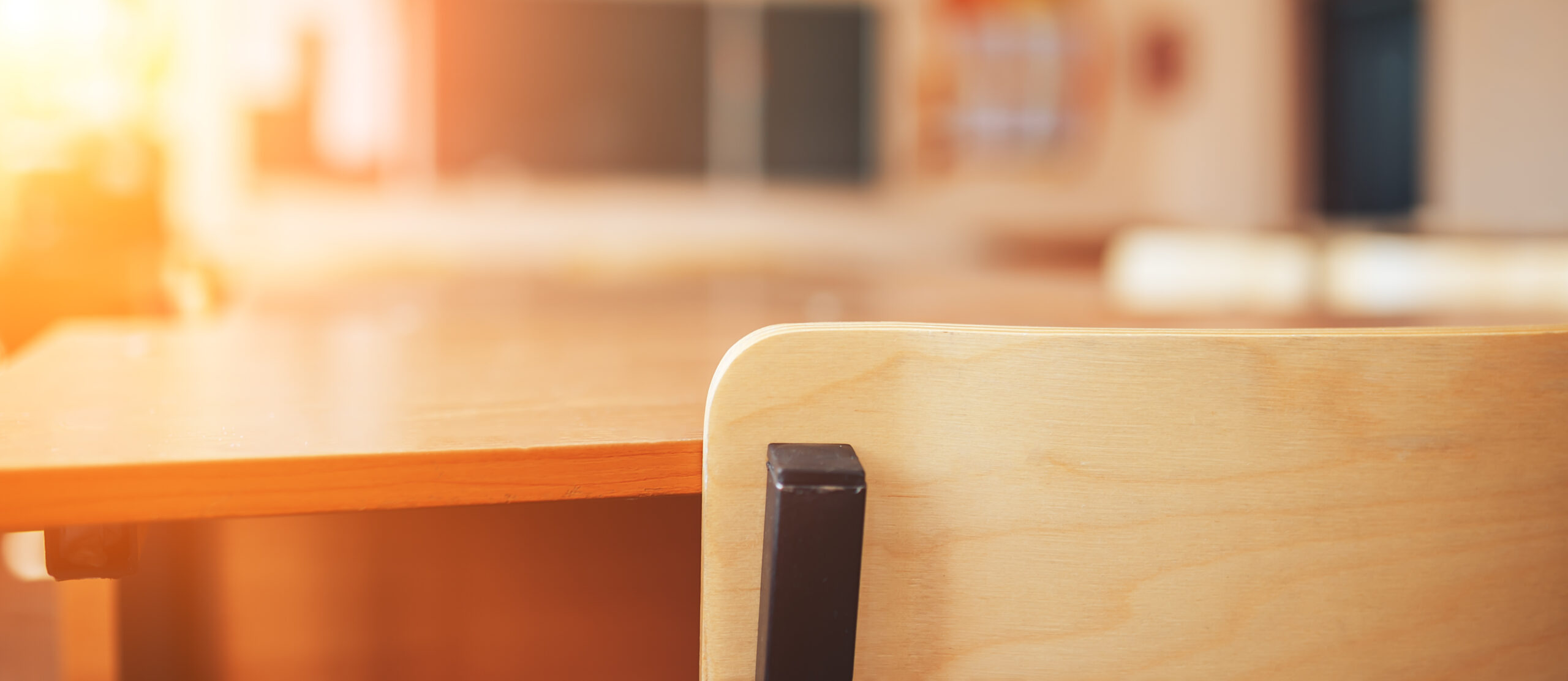 Close-up of an empty wooden desk and chair in a sunlit classroom, ideal for Civic Education. A blurry blackboard and shelves fill the background as warm sunlight streams through the window, creating a calm and inviting atmosphere.