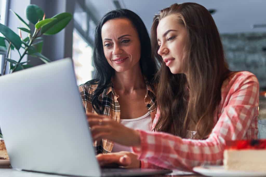 Two women in plaid shirts sit at a table, smiling and looking at a laptop screen together as they explore exciting Youth Programs. There is cake on the table and a plant in the background.