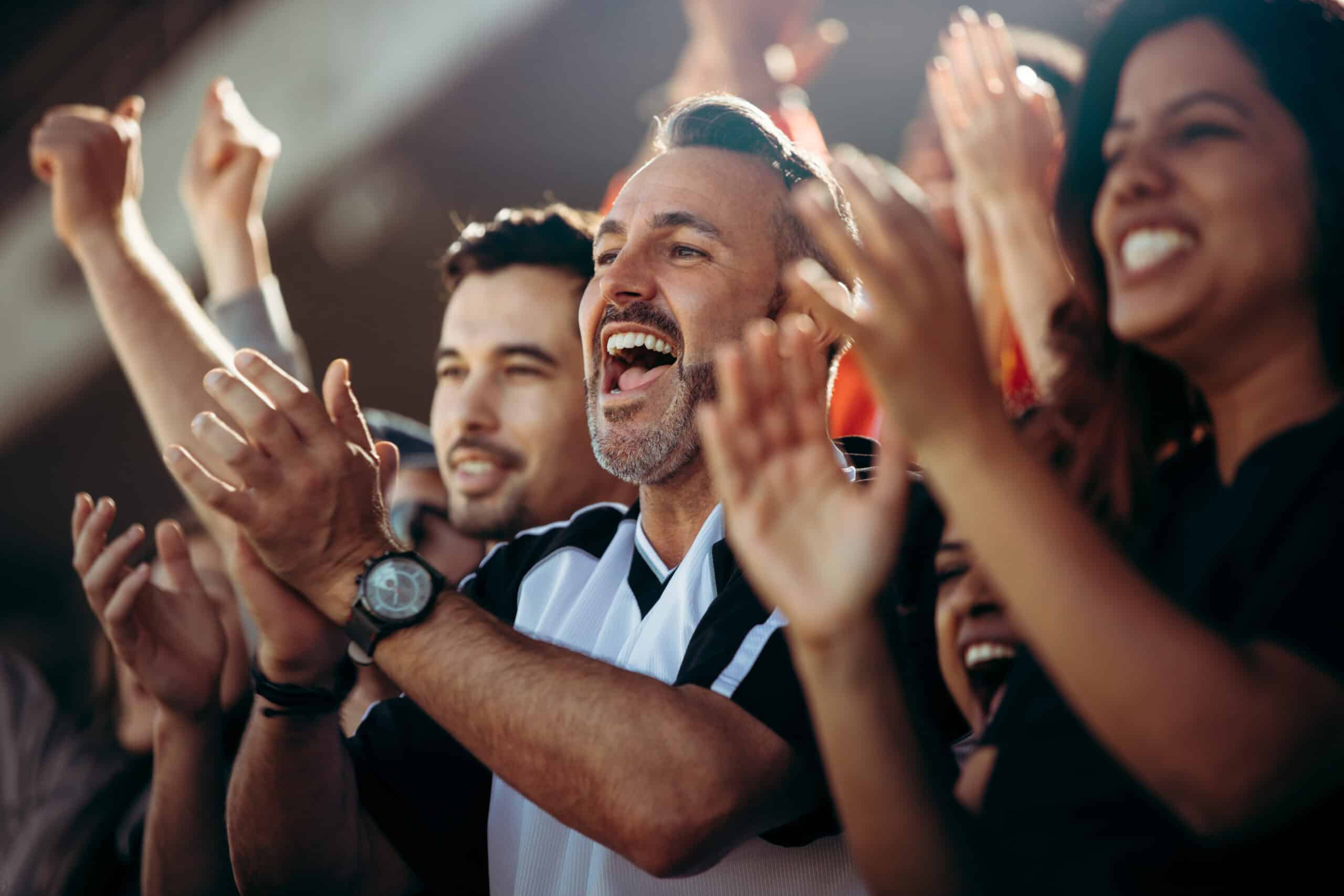 A group of excited fans cheer and clap at a sports event, celebrating the thrilling atmosphere of the World Cup. The focus is on a smiling man in the center wearing a black and white jersey. The crowd around him is energetic and joyful.