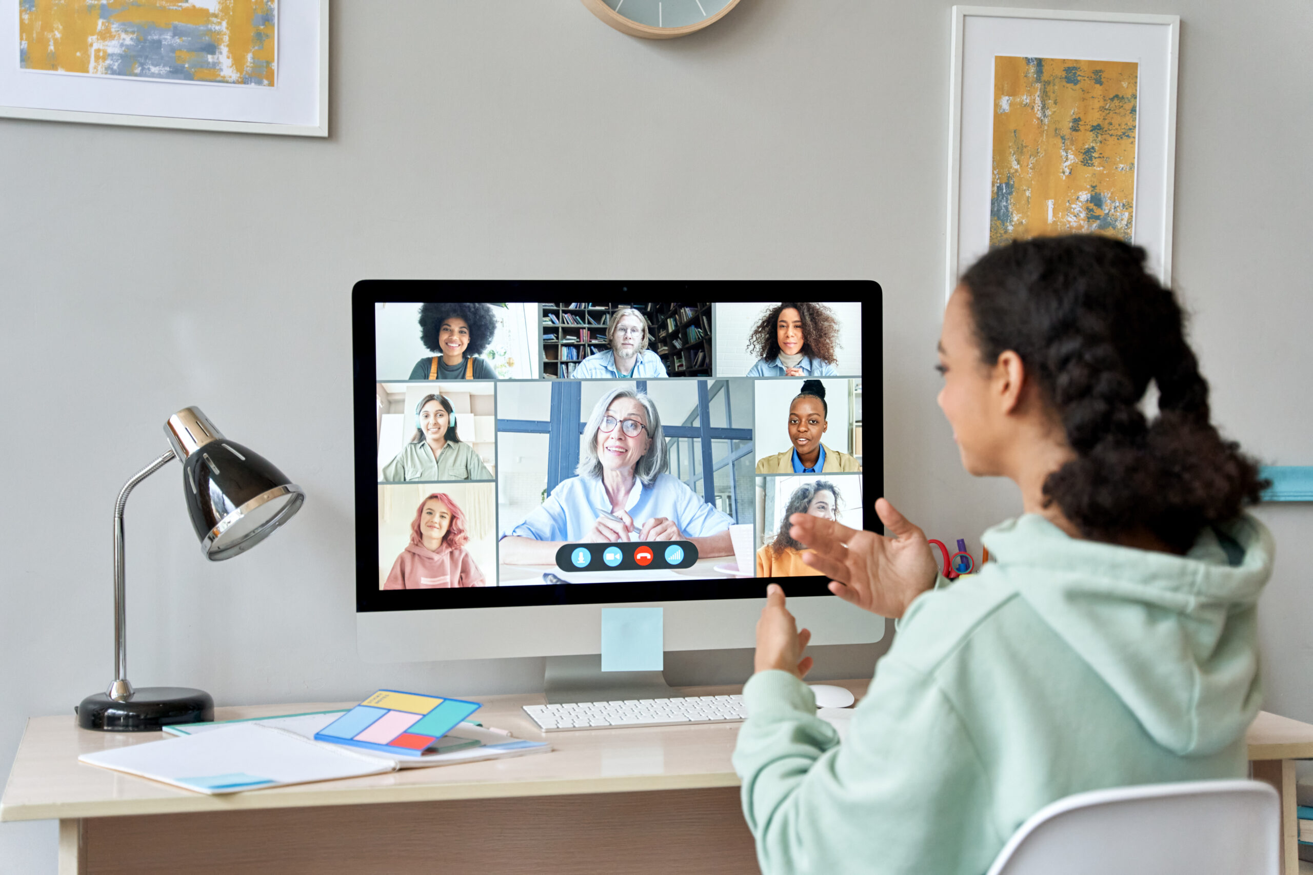 A young woman sits at a desk, joining a video call on her computer with seven diverse ambassadors, all visible in small windows on the monitor. She gestures while speaking, and office supplies are on the desk.