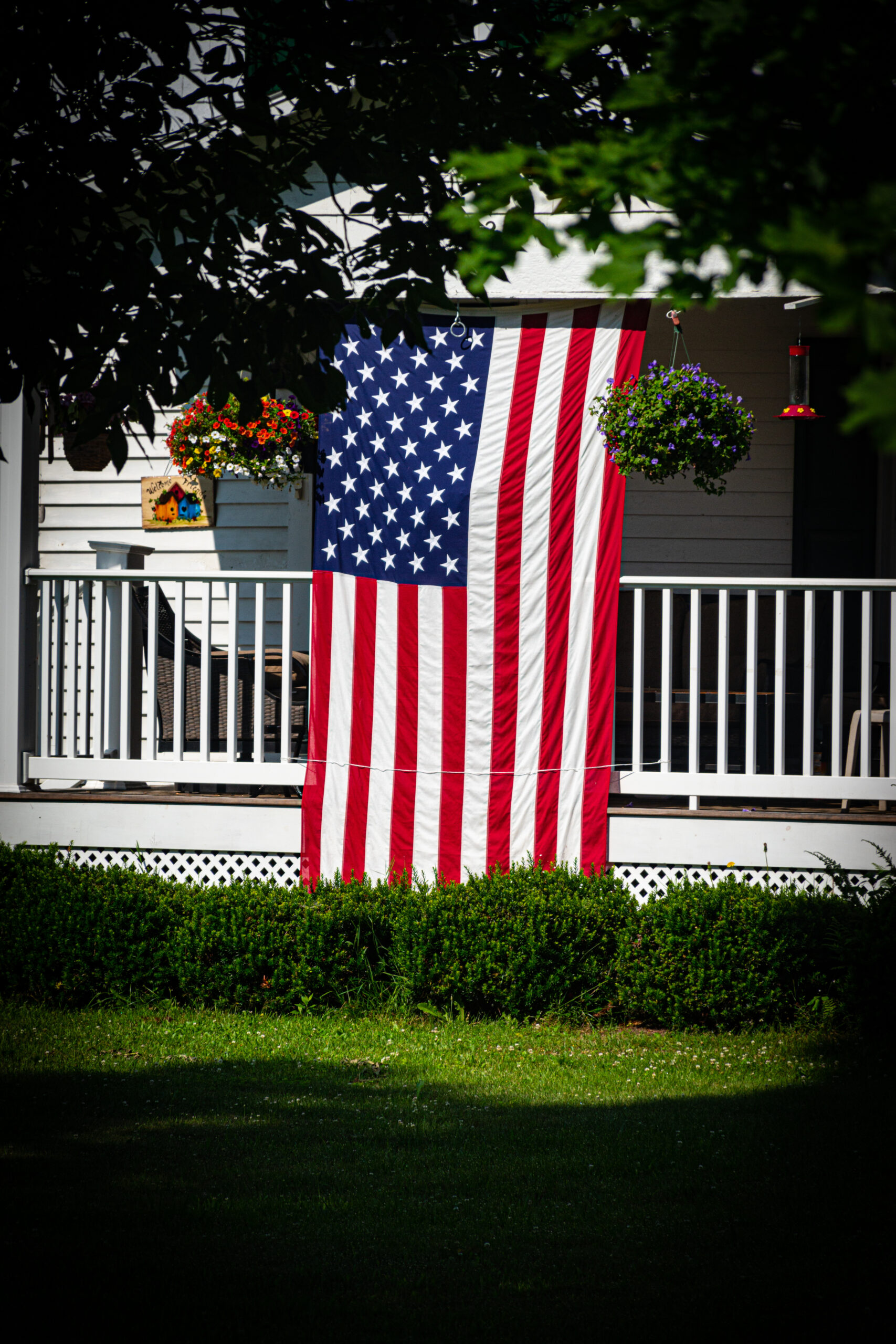 An American flag is hung on the front porch of this home in the small town of Windsor in Broome County in Upstate NY. Proud to fly the Stars and Stripes for all Patriots to see. An upside-down American flag hangs from the porch of a white house with a railing, marking America 250. Flower pots and hanging baskets decorate the porch, while green bushes and lawn are visible in the foreground.