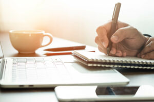 A person writes in a notebook with a pen on a desk, contemplating whether to donate. Nearby, a laptop, smartphone, cup, and eyeglasses rest under soft lighting, creating a warm and focused atmosphere.