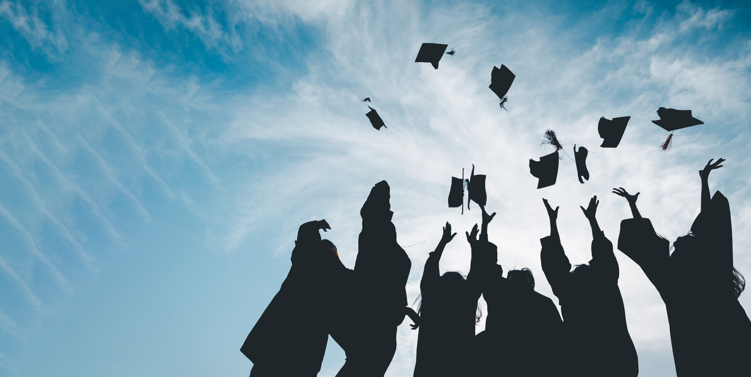 Silhouetted graduates toss their caps into a bright blue sky with wispy clouds, celebrating their achievement.