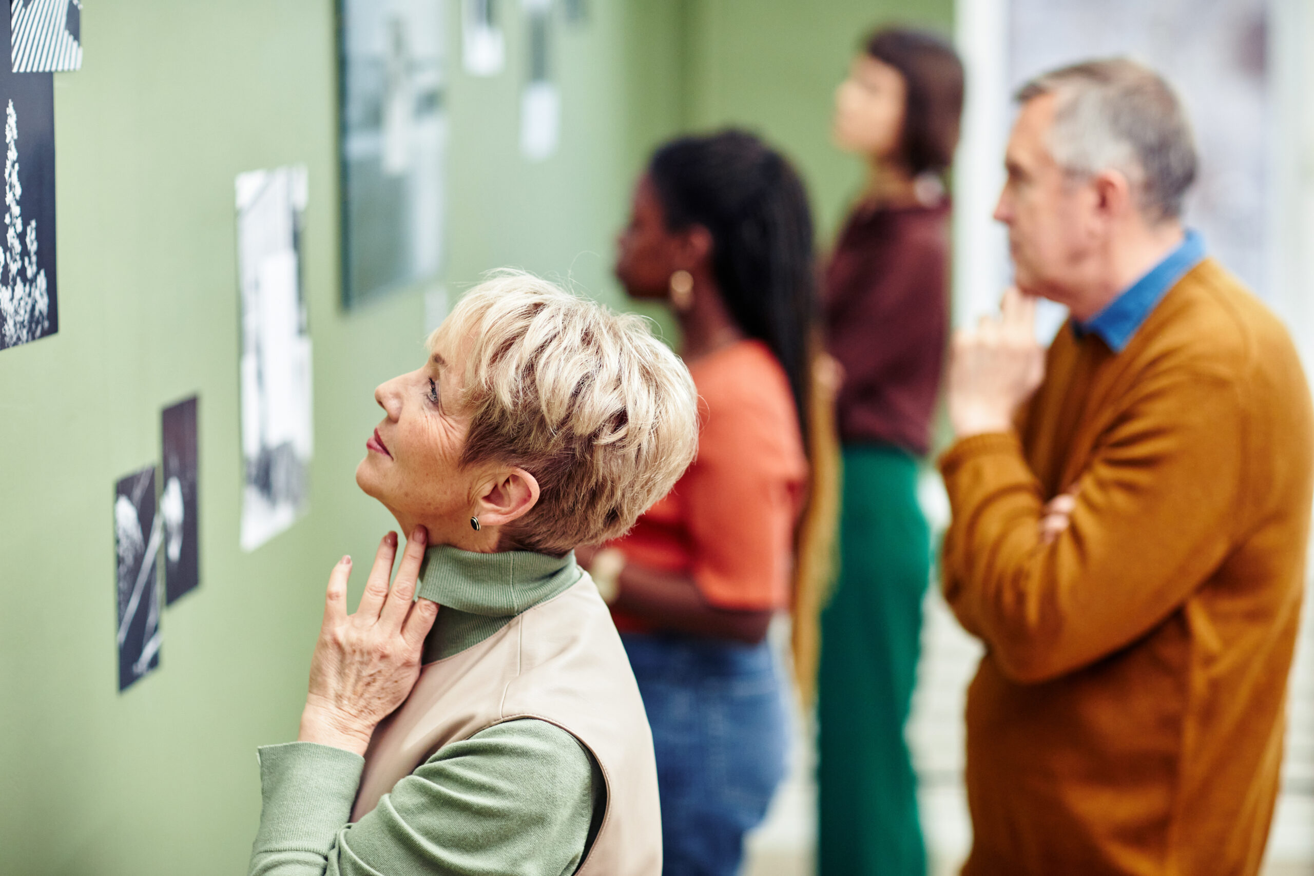 Modern Photography Exhibition Visitors Four adults observe black and white photographs displayed on a green wall in an art gallery. Two people are in focus in the foreground, while two others appreciate the Summer of civics themed photos in the background.