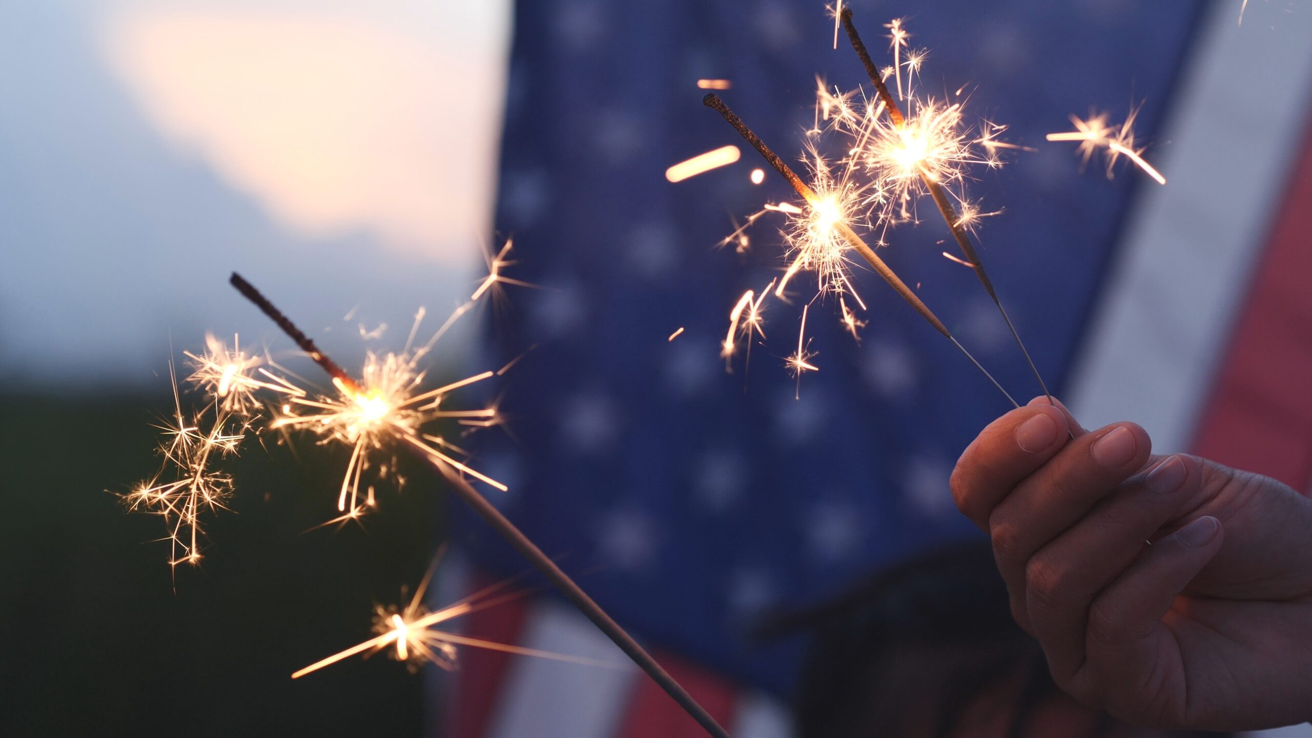 Happy 4th of July Independence Day, Hand holding Sparkler fireworks USA celebration with American flag background. Concept of Fourth of July, Independence Day, Fireworks, Sparkler, Memorial, Veterans A hand holding sparklers, glowing brightly, with a blurred American flag in the background. The scene suggests a festive celebration of America 250, possibly on the Fourth of July.
