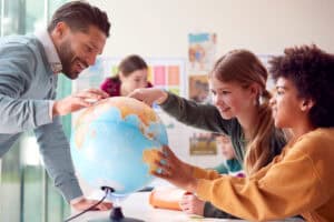 A teacher and two students smile and point at a globe during a spring-themed geography lesson in the classroom. Other students are blurred in the background, with colorful posters and bright spring decorations on the walls.