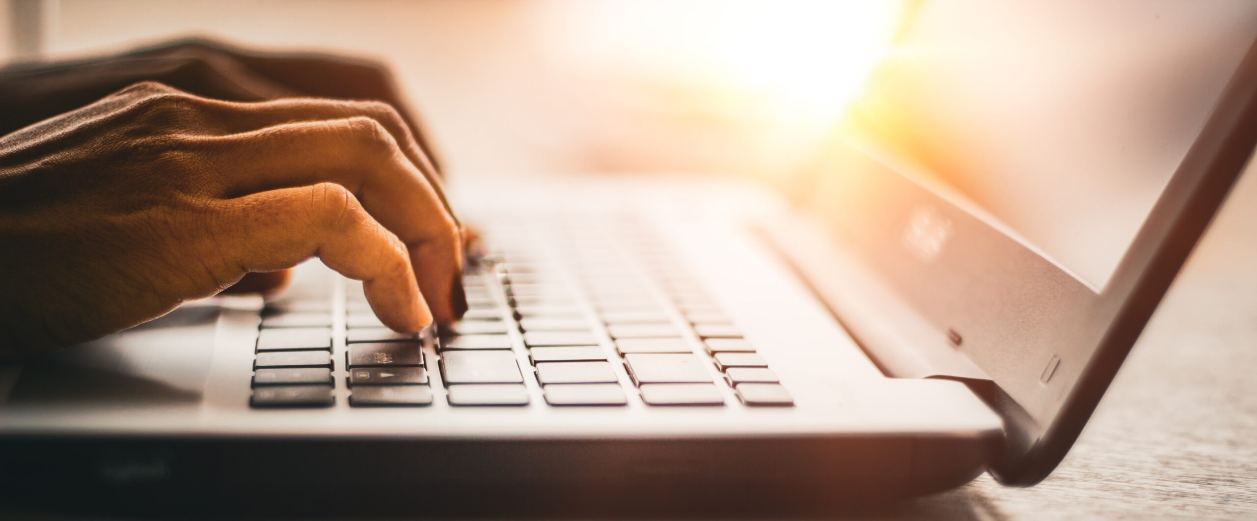 AdobeStock_511434500 Close-up of hands typing on a laptop keyboard with bright sunlight shining in the background, creating a warm and focused atmosphere.