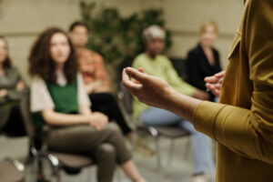 A person in a yellow shirt speaks to a seated group of four, fostering student engagement as they listen attentively in a casual, indoor setting. The focus is on the speaker’s hand in the foreground.