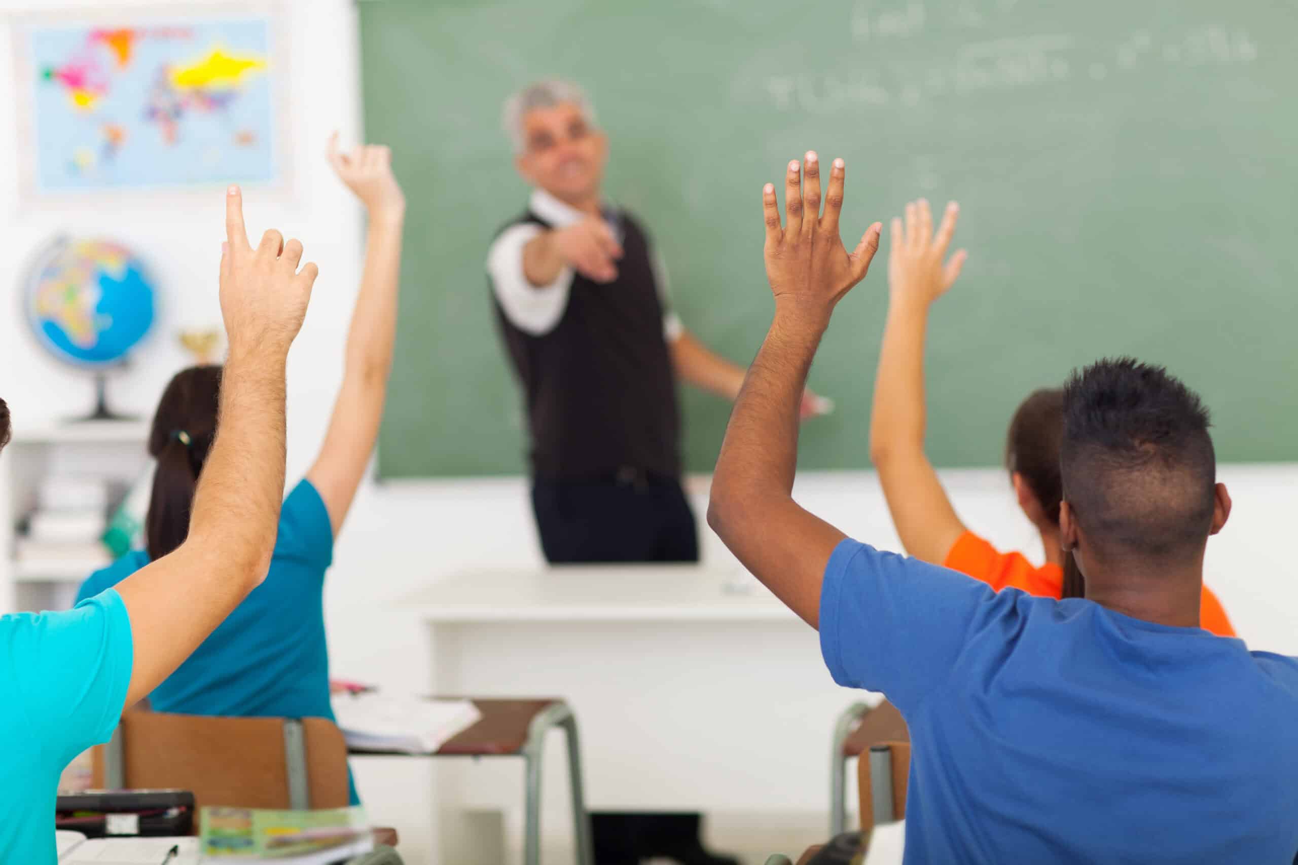 Students sitting at desks raise their hands in a classroom, while a teacher stands at the front near a chalkboard, pointing towards them—prompting an important question: Why Are Students Self-Censoring in Civics Class?.