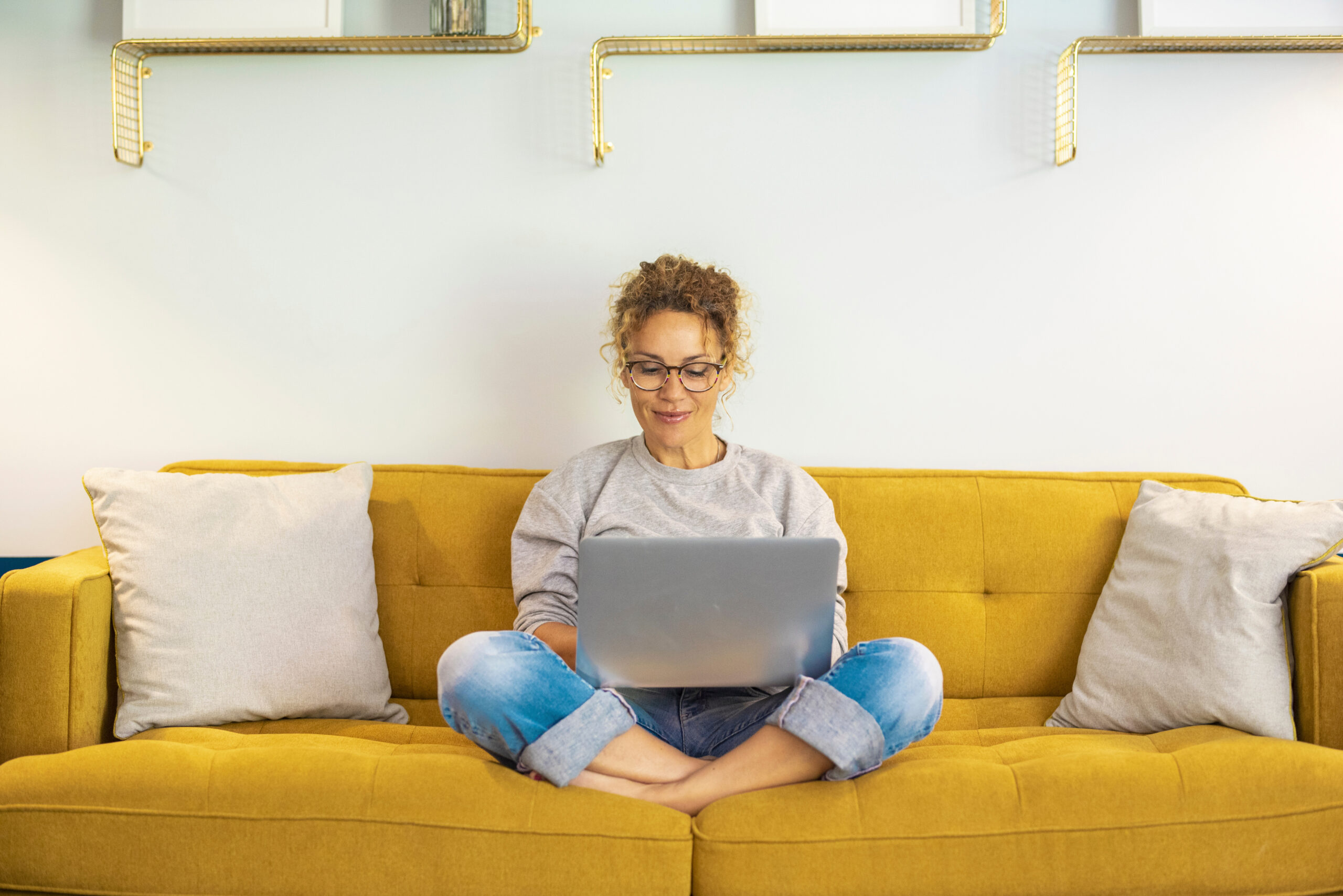 A woman sits cross-legged on a yellow sofa, using a laptop on her lap. With glasses and casual attire, she looks inspired—perhaps reading about Sandra Day O'Connor—with two light pillows beside her and shelves adorning the wall behind.