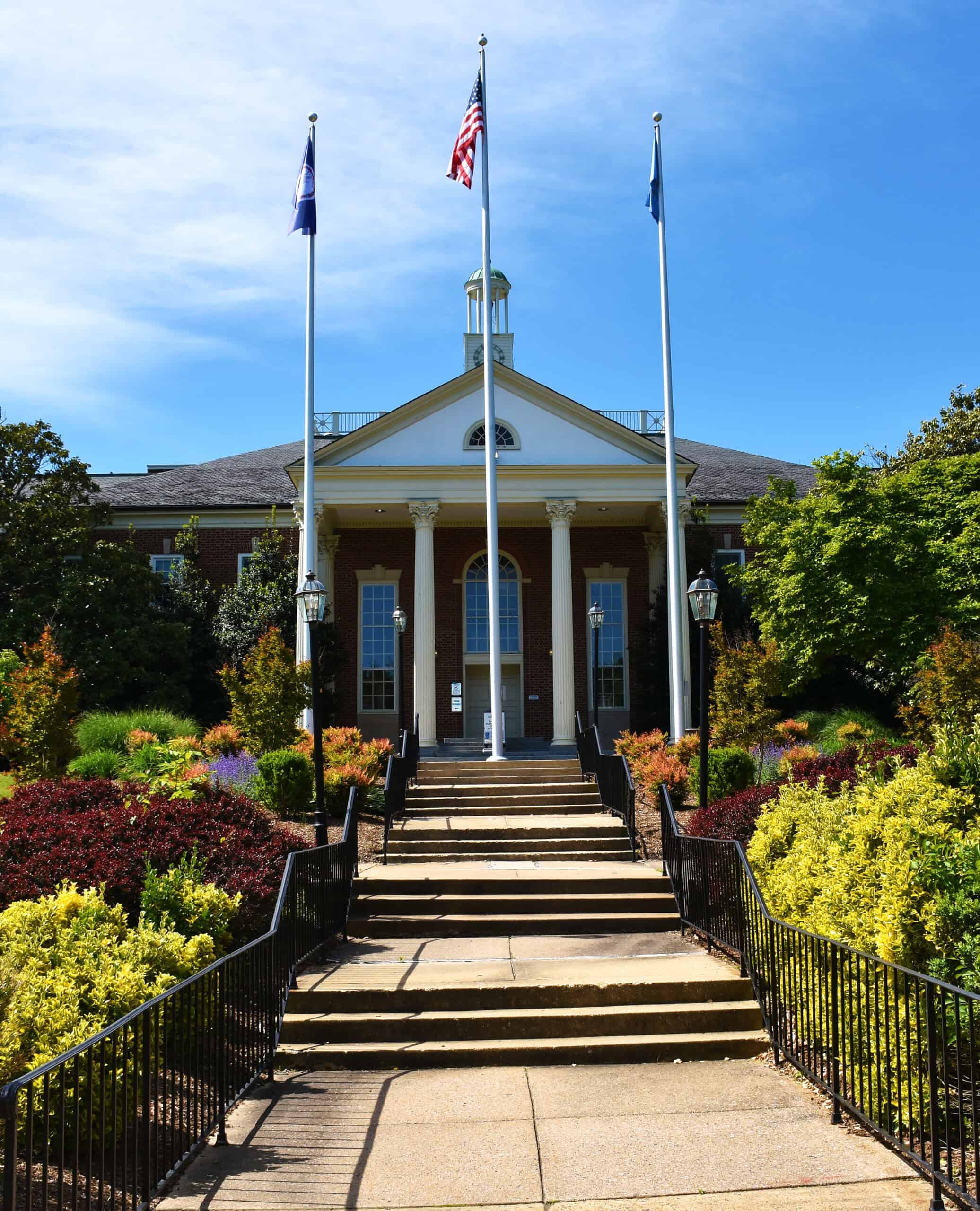 A symmetrical brick building with white columns and three flagpoles stands behind a staircase, surrounded by colorful bushes and greenery under a clear blue sky, capturing the spirit of the Summer of Civics.