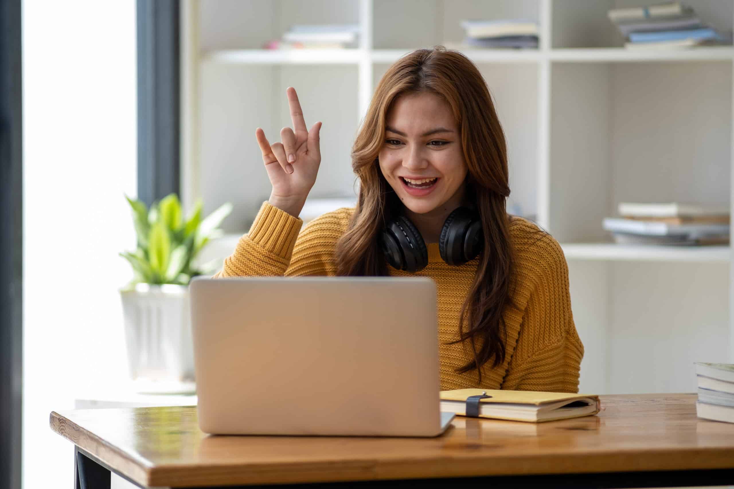 Young woman in a yellow sweater with headphones around her neck smiles and raises her hand while looking at a laptop on a desk, embodying the enthusiasm of modern Ambassadors. Books and a plant are in the background.