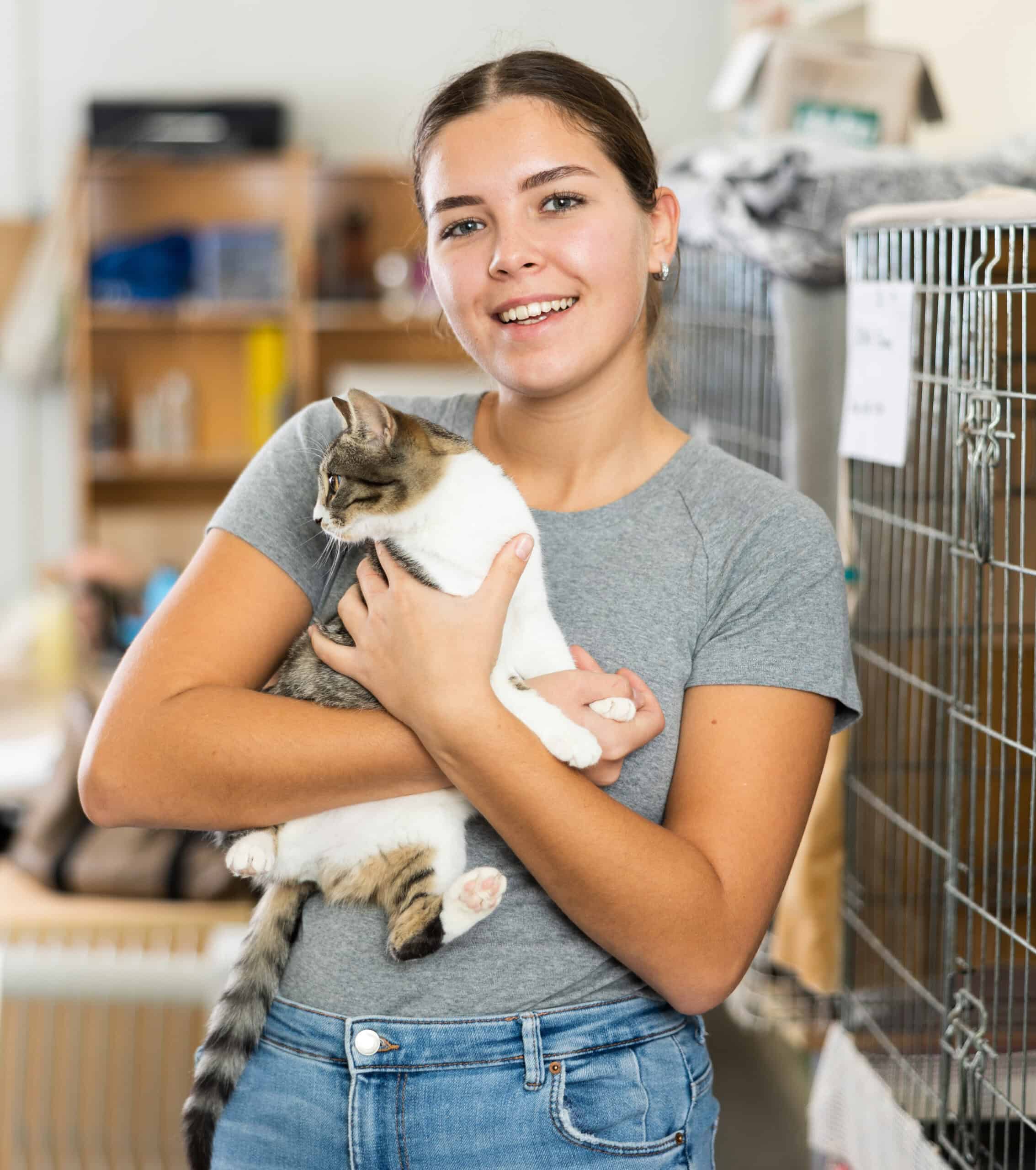 Young woman in a gray shirt smiles while holding a gray and white cat in an animal shelter, with cages and supplies visible in the background—a heartwarming scene reminiscent of the dedication celebrated by the Arizona Community Service Awards.