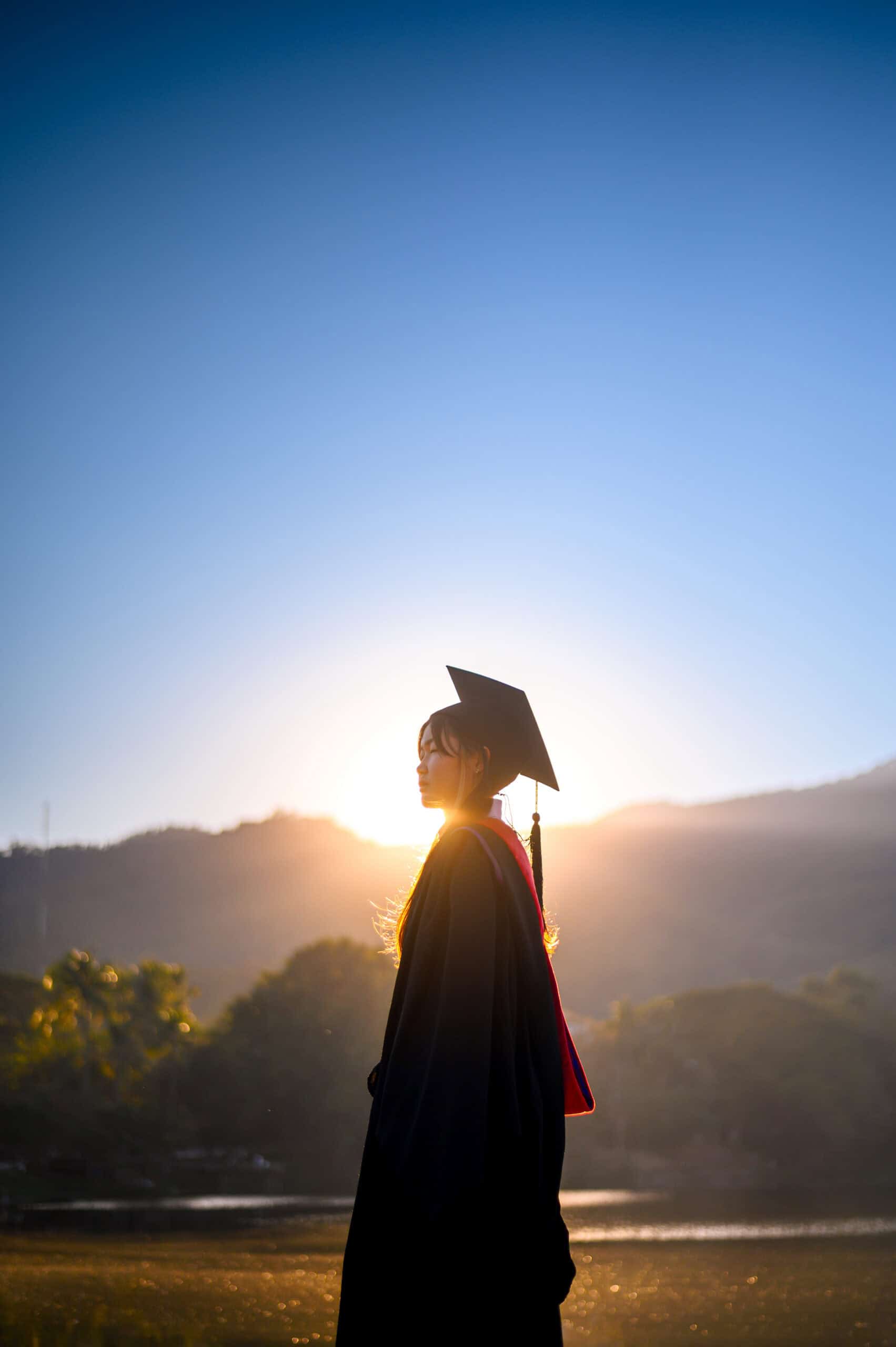 A graduate in a cap and gown stands outdoors at sunrise, embodying The Citizen, with mountains and trees in the background and sunlight glowing behind them.