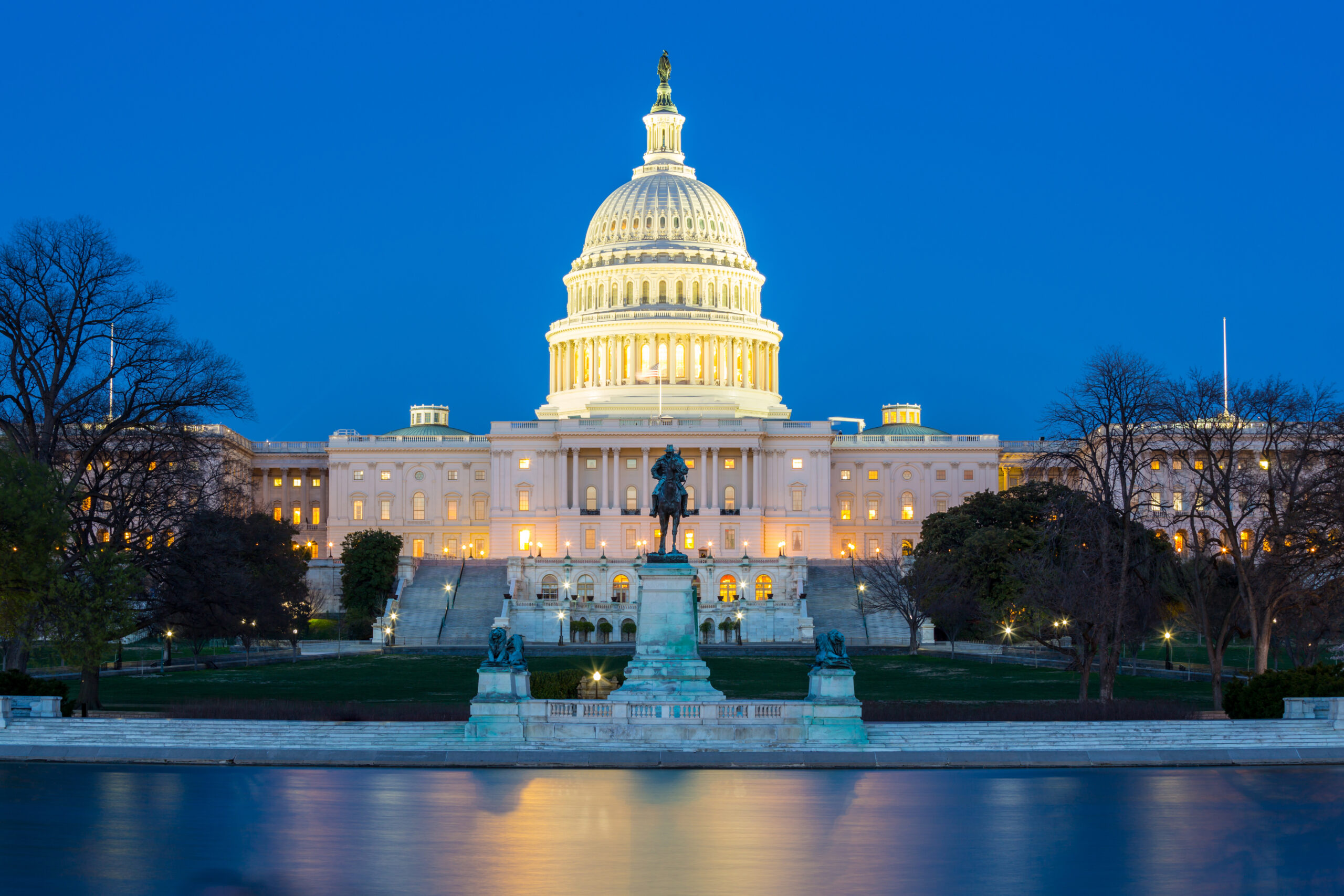 The United States Capitol building illuminated at dusk, with a clear sky backdrop. The dome is prominently lit, and its reflection shimmers in the pool in the foreground. Leafless trees flank the building, indicating a winter setting.