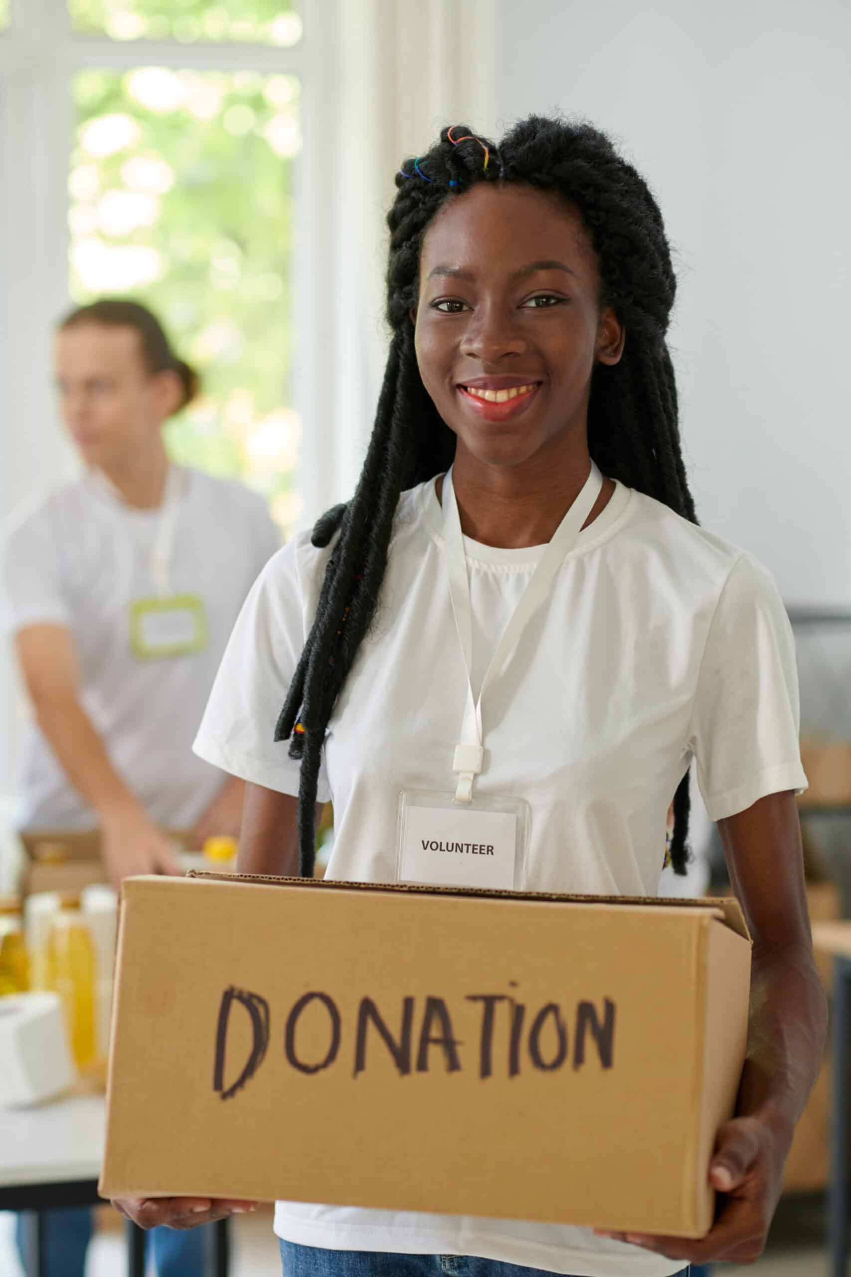 A smiling volunteer in a white shirt holds a cardboard box labeled "DONATION." Another person and food items are visible in the background, reflecting the spirit of giving celebrated by the Arizona Community Service Awards.