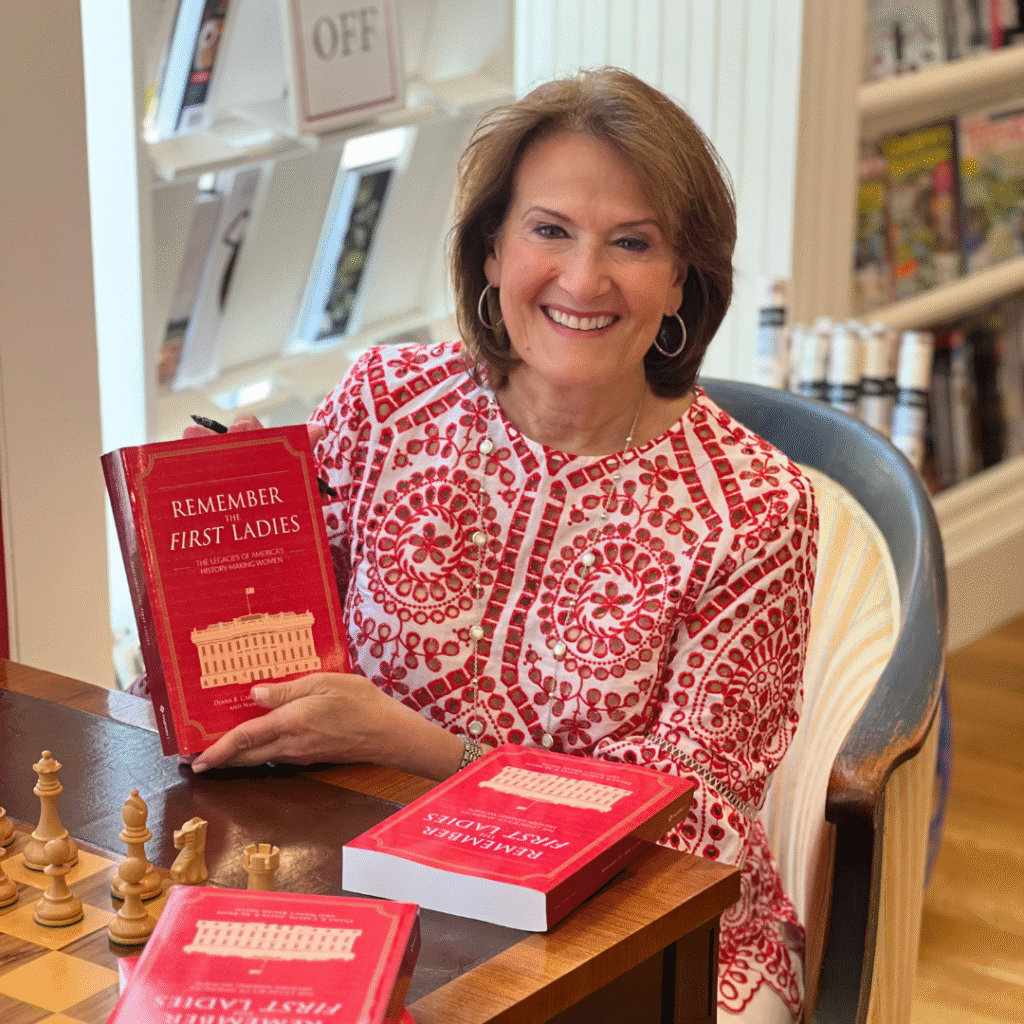 A woman smiling and sitting at a table with chess pieces, holding a red book titled "Remember the First Ladies," celebrates women in leadership. More copies of the book are on the table, with bookshelves and magazines in the background.