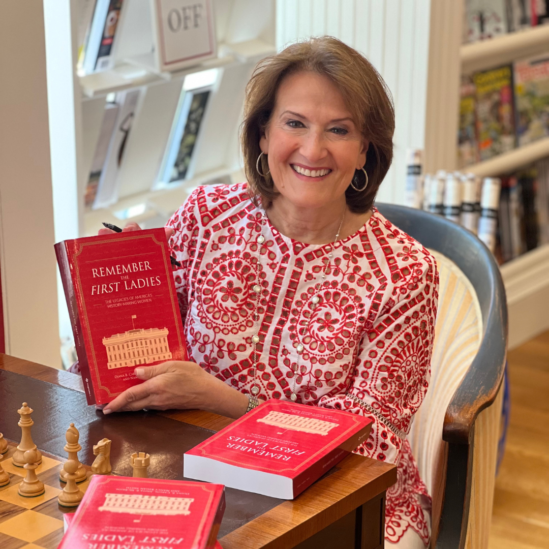 A woman smiling and sitting at a table with chess pieces, holding a red book titled "Remember the First Ladies," celebrates women in leadership. More copies of the book are on the table, with bookshelves and magazines in the background.