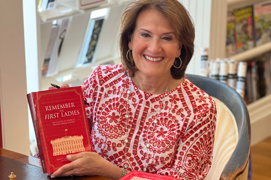 A woman in a red and white patterned blouse smiles while holding up a book titled "Remember the First Ladies," celebrating women in leadership. She is seated at a table with bookshelves and magazines in the background.