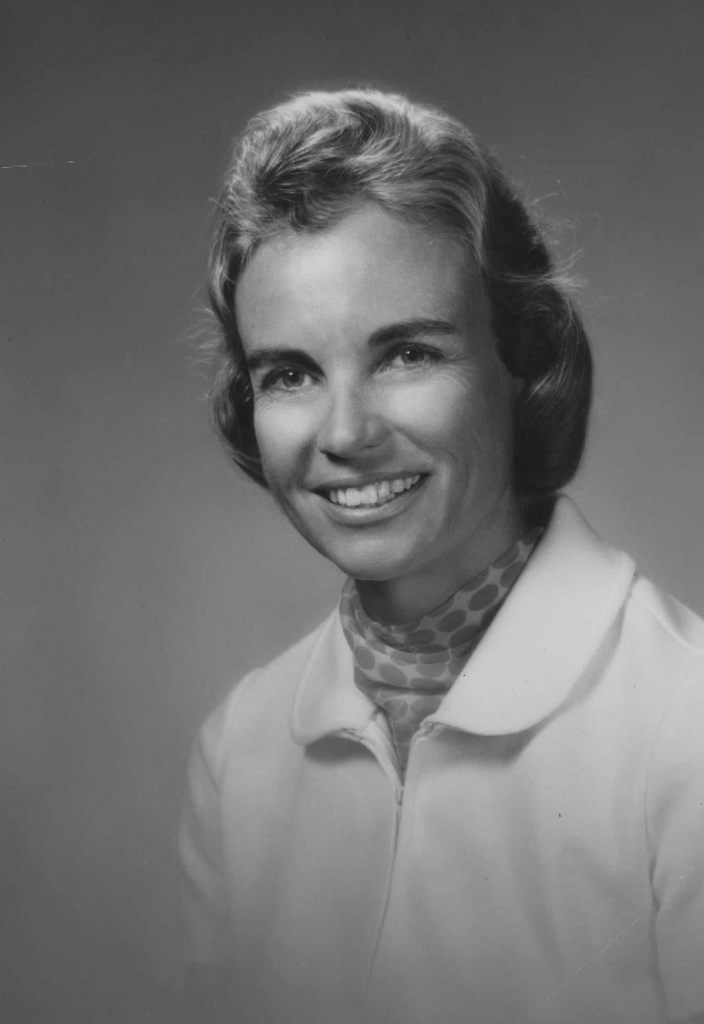 Black and white photo of a smiling woman with short, wavy hair, wearing a light-colored jacket and a patterned scarf around her neck. She looks directly at the camera against a plain background.