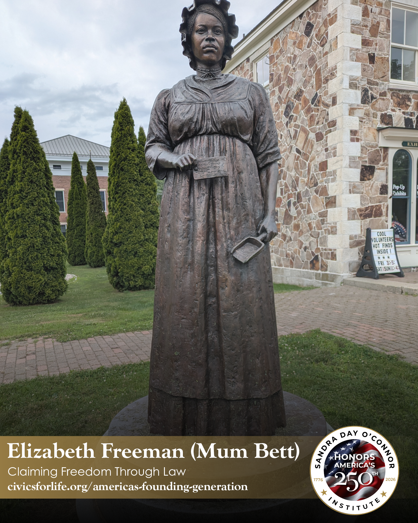 A bronze statue of Elizabeth Freeman (Mum Bett), known as The Citizen, stands outdoors on a brick walkway, with tall green shrubs and a stone building with large windows in the background.